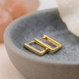 Könnte beinhalten: Ein Paar kleiner, goldfarbener, rechteckiger Creolen mit polierter Oberfläche. Die Ohrringe verfügen über einen Klappverschluss und eine klare, geometrische Form und liegen als Set mit einer glatten, reflektierenden Metalloberfläche.