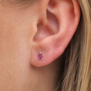 Puede incluir: Primer plano de un pendiente de plata con una gema rosa brillante en forma de lágrima. El pendiente está engastado en un bisel de plata y se lleva en una oreja. Una joya sencilla y elegante.
