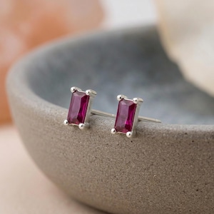 May include: A pair of stud earrings featuring rectangular baguette-cut deep pink gemstones set in silver-toned metal. Each earring has four small prongs securing the stone, with a straight post extending from the back of the setting.