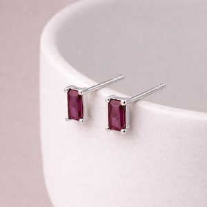 Puede incluir: Un par de pendientes de plata con gemas rectangulares de color rojo intenso. Los pendientes están engastados en un bisel de plata y tienen un diseño sencillo y elegante. Los pendientes se muestran sobre una superficie blanca.