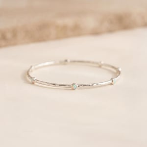 Pode incluir: Um anel fino de metal prateado com uma superfície martelada e texturizada. A banda é decorada com várias pedras pequenas, redondas e iridescentes semelhantes a opalas, colocadas a intervalos regulares ao redor de todo o anel.