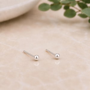 Pode incluir: Dois brincos de botão prateados com um pequeno design de bola redonda. Os brincos são exibidos em uma superfície clara e texturizada. Os brincos são simples e elegantes, adequados para uso diário.
