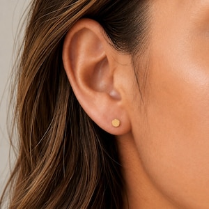 May include: A small, circular gold stud earring with a smooth, polished finish. The earring is positioned in the centre of the earlobe, featuring a simple, flat, round design that sits flush against the skin, providing a subtle and clean look.