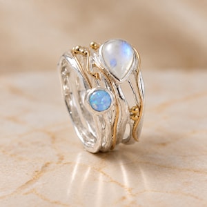 May include: A multi-band ring crafted from silver and gold-toned metal, featuring a pear-shaped moonstone and a round, iridescent blue opal. The design incorporates organic, flowing lines and small metallic spheres, creating a layered appearance.