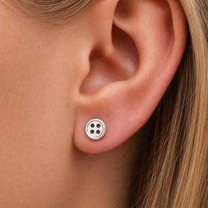 May include: A silver button stud earring with four small holes is shown on an ear. The earring is round and has a simple, classic design. The background is a person's skin and hair.