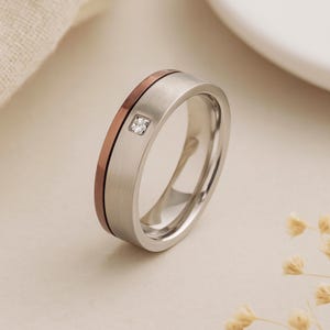 Peut inclure: Une bague argentée avec une finition en métal brossé et une bande de couleur cuivre. La bague comporte une petite pierre précieuse transparente de coupe carrée. C'est un bijou.
