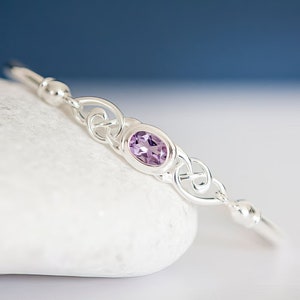 May include: A silver bangle bracelet featuring a central oval-cut purple amethyst gemstone set in a polished metal frame. The design includes intricate silver knotwork detailing on both sides of the stone, flanked by smooth silver beads.
