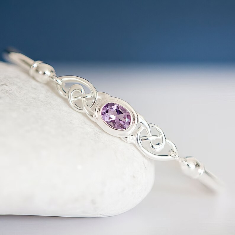 May include: A silver bangle bracelet featuring a central oval-cut purple amethyst gemstone set in a polished metal frame. The design includes intricate silver knotwork detailing on both sides of the stone, flanked by smooth silver beads.