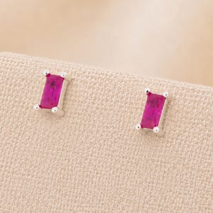Könnte beinhalten: Ein Paar Ohrstecker mit rechteckigen, im Baguetteschliff geschliffenen, leuchtend pinken Edelsteinen in poliertem silberfarbenem Metall. Jeder Ohrstecker ist mit vier kleinen Krappen an den Ecken befestigt, was ein klares, geometrisches Design ergibt.