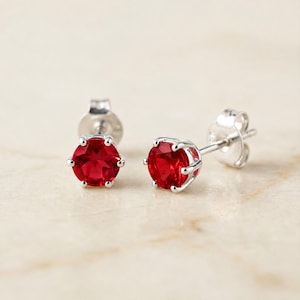 May include: A pair of stud earrings featuring round, faceted, vibrant red stones set in polished silver-toned metal. Each earring is secured with a matching silver-toned post and butterfly back, showcasing a classic six-prong setting design.