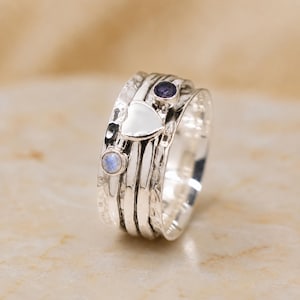 May include: A wide silver spinner ring featuring a hammered texture, a central polished silver heart charm, and two small round bezel-set gemstones, one dark purple and one iridescent white, mounted on the rotating outer band of the ring.