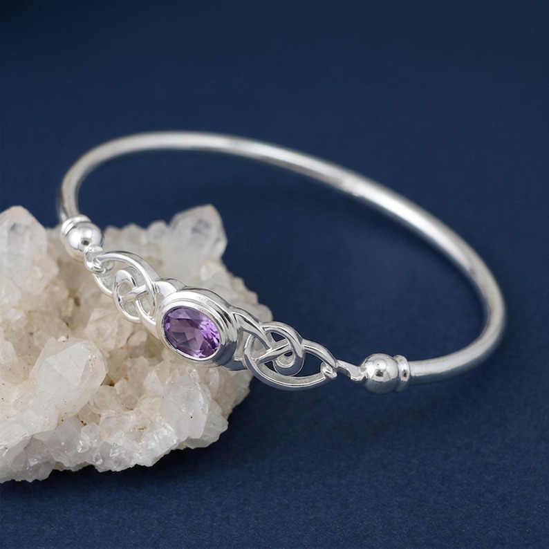 May include: A silver bangle bracelet featuring a central oval-shaped purple amethyst gemstone. The metal band displays intricate knotwork detailing on both sides of the stone, finished with smooth silver spheres at the connection points.