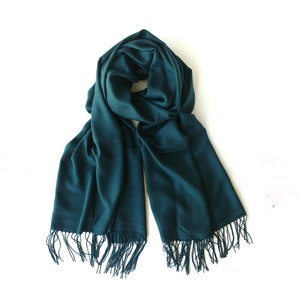 Puede incluir: Una bufanda azul turquesa con flecos en los bordes. La bufanda está doblada por la mitad y colocada sobre un fondo blanco.