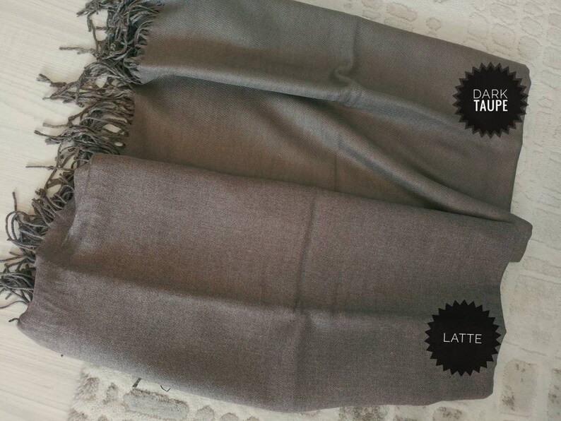 Puede incluir: Una suave bufanda marr&oacute;n con flecos en dos tonos de marr&oacute;n, etiquetada como "Dark Taupe" y "Latte".