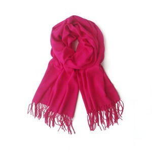 May include: A bright pink scarf with fringe on the ends. The scarf is folded in half and draped over a white surface.