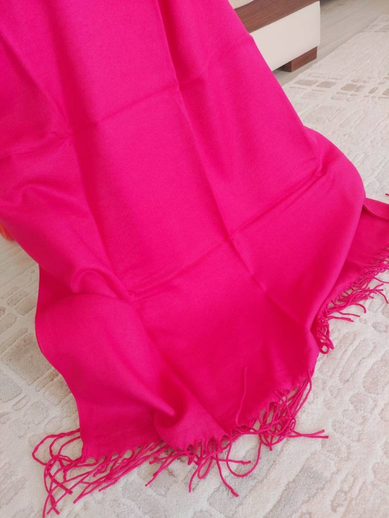 May include: A bright pink shawl with fringe detailing. The shawl is made of a soft, lightweight fabric.
