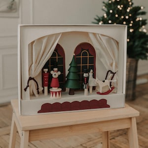 Op de afbeelding: Een houten kerstscène met een kleine boom, een notenkraker, een beer en een schommelpaard. De scène is geplaatst in een witte doos met een rood en wit gordijn.