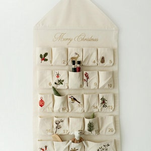 Può includere: Un calendario dell'avvento in tessuto color crema con una parte superiore a punta e la scritta Merry Christmas. Presenta ventiquattro tasche individuali, ciascuna numerata e decorata con motivi botanici o natalizi ricamati, tra cui un cervo.