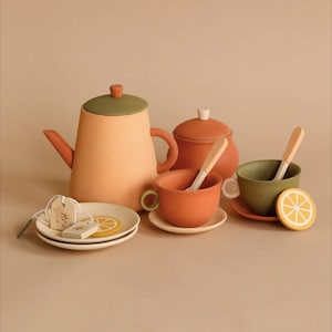 Puede incluir: Un juego de té de madera con una tetera, un azucarero, dos tazas, dos platillos y dos cucharas. La tetera y el azucarero son de color terracota con una tapa verde. Las tazas son de color terracota y los platillos son de color marrón claro. Las cucharas son de color marrón claro. Hay dos rodajas de limón en los platillos.