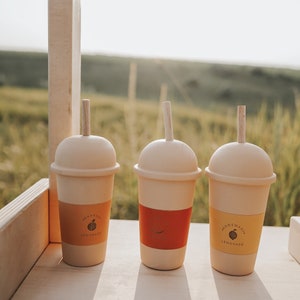 May include: Three light-colored wooden toy cups with dome lids and vertical wooden straws. Each cup features a colored paper sleeve with printed text and fruit illustrations, including orange, watermelon, and lemon designs with the word lemonade.