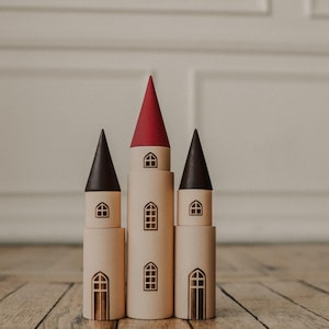 Peut inclure: Château jouet en bois avec trois tours. Les tours ont des toits coniques marron foncé et rouges. Chaque tour comporte de petites découpes de fenêtres. Le château est en bois de couleur claire et repose sur une surface en bois.