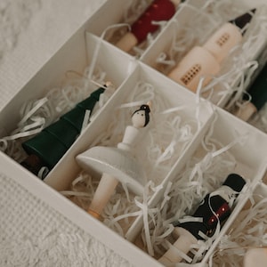 May include: A white box filled with white shredded paper and various wooden Christmas decorations. The decorations include a ballerina, a Christmas tree, a soldier, a church, and a star.