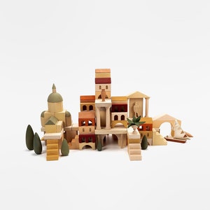 May include: A wooden toy set featuring a miniature town with buildings, trees, and a bridge. The buildings are made of stacked blocks in various shades of brown, yellow, and red. The set includes a small boat and a figure of a person.