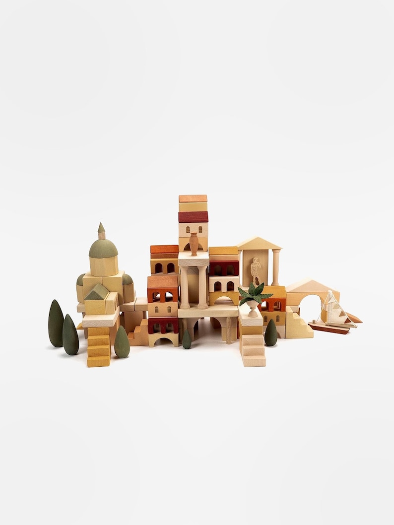 May include: A wooden toy set featuring a miniature town with buildings, trees, and a bridge. The buildings are made of stacked blocks in various shades of brown, yellow, and red. The set includes a small boat and a figure of a person.