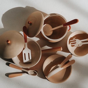 Könnte beinhalten: Ein Set aus hölzernen Spielküchenutensilien und Geschirr in Beige- und Orangetönen. Das Set enthält einen Topf mit Deckel, einen Spatel, einen Löffel, eine Gabel und zwei Schüsseln.