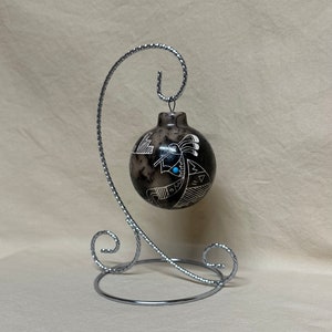 May include: A black and white hand-painted ceramic ornament with a Native American design. The ornament is on a silver stand.