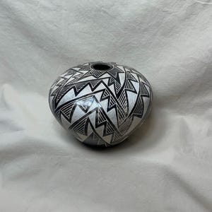 May include: A round, ceramic vase with a black and white geometric pattern. The vase has a small opening at the top and is decorated with a repeating zigzag design. The color scheme is black and white.