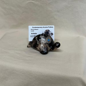 Puede incluir: Figura de tortuga de cerámica con un acabado oscuro y moteado y diseños tribales blancos. La tortuga tiene un ojo de color turquesa y una pequeña tarjeta de visita detrás que dice "Contemporary Acoma Pottery".
