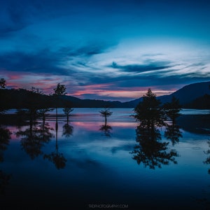 May include: A serene landscape photograph of a lake at dusk. The sky transitions from deep blue to pink and purple hues, reflecting in the still water. Silhouetted trees stand in the water, mirroring their forms. Mountains are visible in the background.