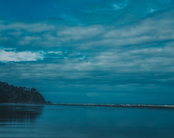 May include: A panoramic view of a beach with a dark blue sky and a line of trees in the distance. The water is calm and reflects the sky.