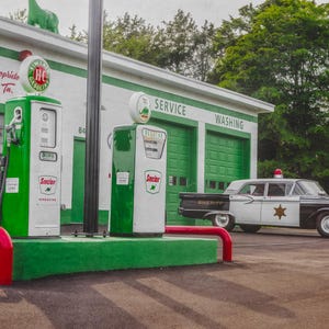May include: A vintage Sinclair gas station with two green and white fuel pumps, a white building with green trim labeled Service and Washing, and a black and white sheriff patrol car parked in front of the garage bays under a cloudy sky.