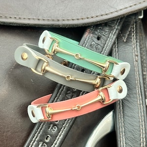 May include: Three leather bracelets with gold-toned equestrian-style snaffle bit charms. The bracelets are in shades of green, grey, and pink.