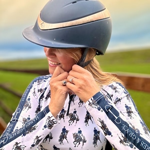 May include: A woman wearing a white long-sleeved shirt with a repeating pattern of horses and riders. The shirt has black trim and the text "HARES & GRACE" on the sleeves. She is wearing a dark blue riding helmet with a gold trim.