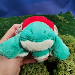 Puede incluir: Un peluche de tortuga con cabeza y aletas turquesas, vientre blanco y caparazón rojo. El juguete está hecho de material suave y difuso y tiene una expresión amigable. El fondo es un cielo azul y musgo verde.