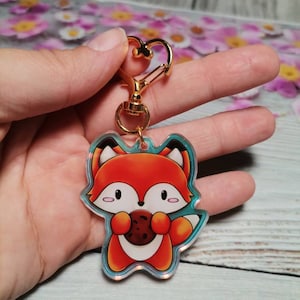 Könnte beinhalten: Ein roter Fuchs-Schlüsselanhänger mit einem goldenen Metallverschluss. Der Fuchs hält einen Schokoladenkeks. Der Schlüsselanhänger ist aus klarem Acryl mit einem holografischen Finish.