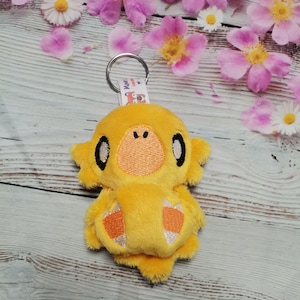 May include: A yellow plush keychain shaped like a bird with orange accents and black eyes. The keychain has a silver metal ring and a white tag that says "Made with Handmade Love".