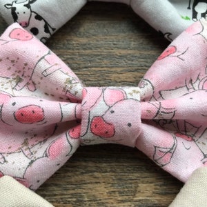 May include: A pink bow tie with a white background and a pattern of pink pigs. The pigs are in various poses, some are standing, some are lying down, and some are looking up.