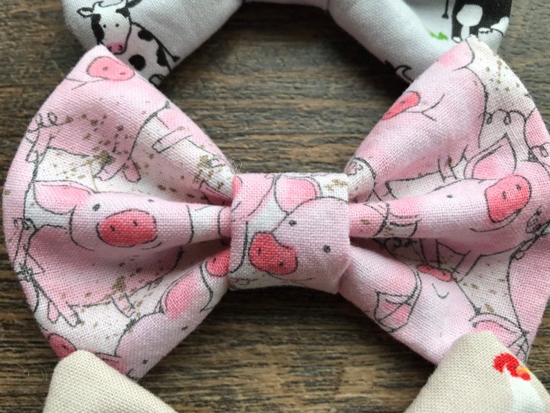 May include: A pink bow tie with a white background and a pattern of pink pigs. The pigs are in various poses, some are standing, some are lying down, and some are looking up.