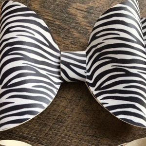 May include: A large, handmade bow with a zebra print pattern in black and white. The bow is made of a stiff material and has a classic bow shape. The bow is likely for hair accessories or gift wrapping.
