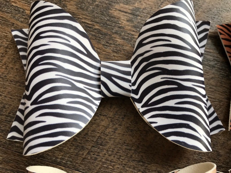 May include: A large, handmade bow with a zebra print pattern in black and white. The bow is made of a stiff material and has a classic bow shape. The bow is likely for hair accessories or gift wrapping.