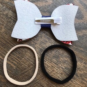 May include: A handmade hair bow with a white felt backing, a navy blue center, and red glitter accents. Includes a metal clip and two hair ties, one beige and one black. Perfect for accessorizing.