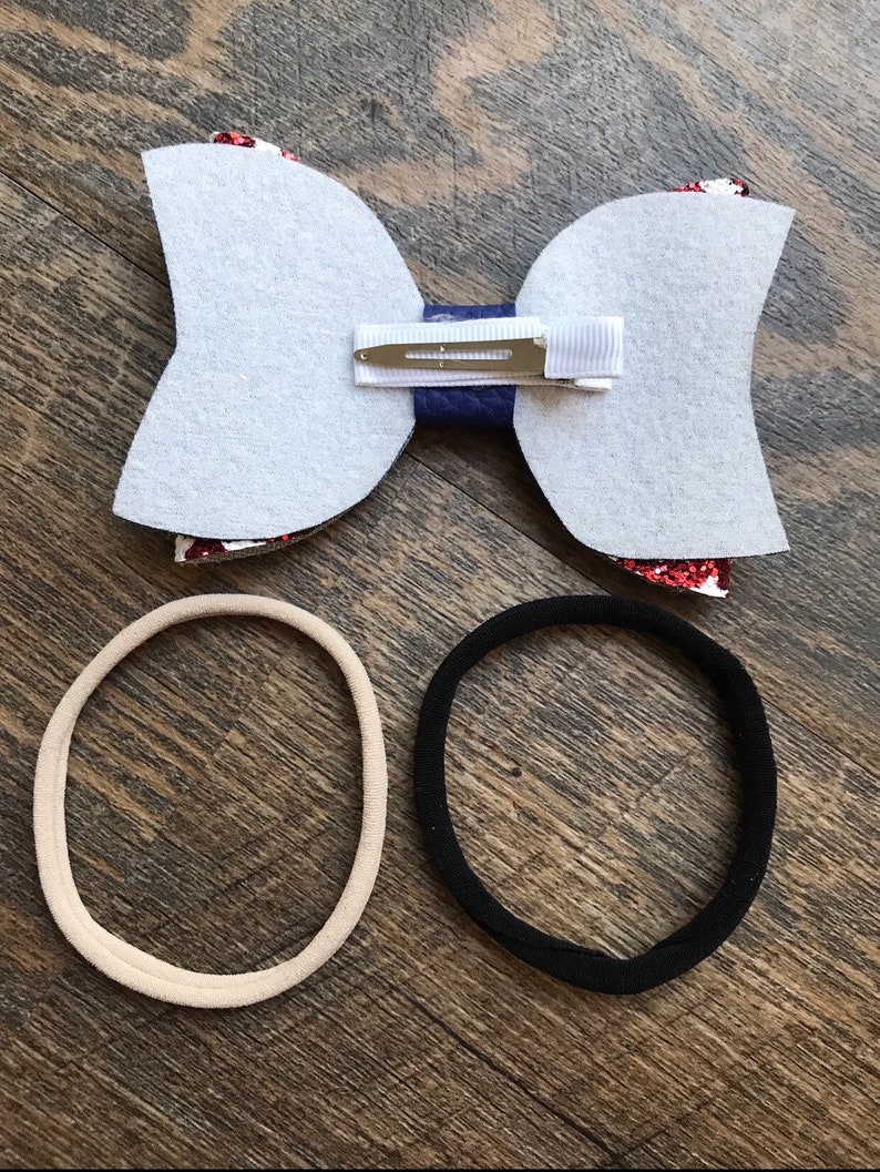 May include: A handmade hair bow with a white felt backing, a navy blue center, and red glitter accents. Includes a metal clip and two hair ties, one beige and one black. Perfect for accessorizing.