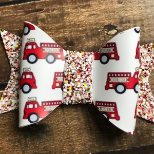 May include: A red and white glitter hair bow with a fire truck pattern. The bow is made of fabric and has a white background with red fire trucks.