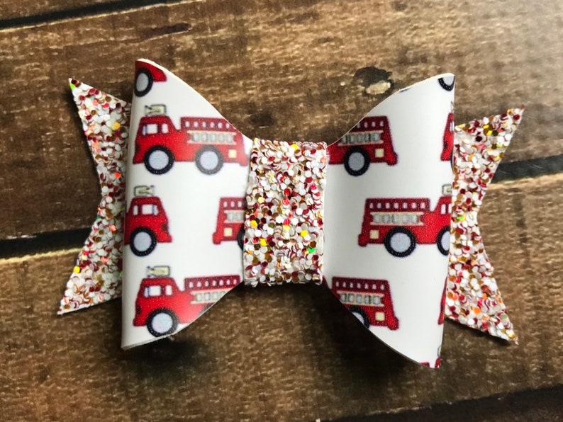 May include: A red and white glitter hair bow with a fire truck pattern. The bow is made of fabric and has a white background with red fire trucks.