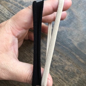 May include: Two hair ties are held up against a wooden surface. One is a flat, black elastic band, and the other is a round, beige elastic hair tie. The hair ties are for styling hair.