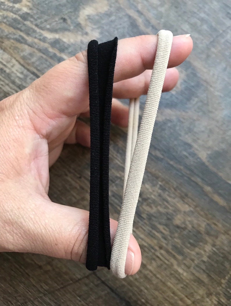 May include: Two hair ties are held up against a wooden surface. One is a flat, black elastic band, and the other is a round, beige elastic hair tie. The hair ties are for styling hair.